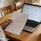 A person sitting at a tidy dining table in an Australian home in daylight reviewing a printed document and an open laptop, seen from the side so the papers and screen are angled away, with a cup of tea on the table.