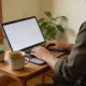 A person typing on an open laptop at a small wooden table in a bright Australian home in the morning, seen from behind with the laptop screen angled away, a mug of tea and a small potted plant beside it in soft window light.
