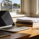 A tidy home-office desk by a window in an Australian house in warm afternoon light, with an open laptop screen angled away and blank, a closed notebook, a stack of books and printed papers, and a cup of tea.