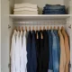 An open wardrobe with a neat row of clothes on wooden hangers in a calm neutral palette of white, navy, black, denim and sand, with folded knitwear and jeans on the shelf above, in a bright Australian bedroom.