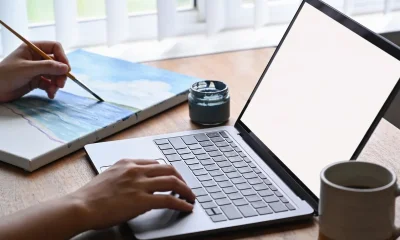 A person's hands holding a paintbrush over a canvas at a desk in a bright Australian home studio, with the other hand resting on a laptop keyboard, a pot of paint and a coffee mug nearby.