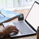 A person's hands holding a paintbrush over a canvas at a desk in a bright Australian home studio, with the other hand resting on a laptop keyboard, a pot of paint and a coffee mug nearby.