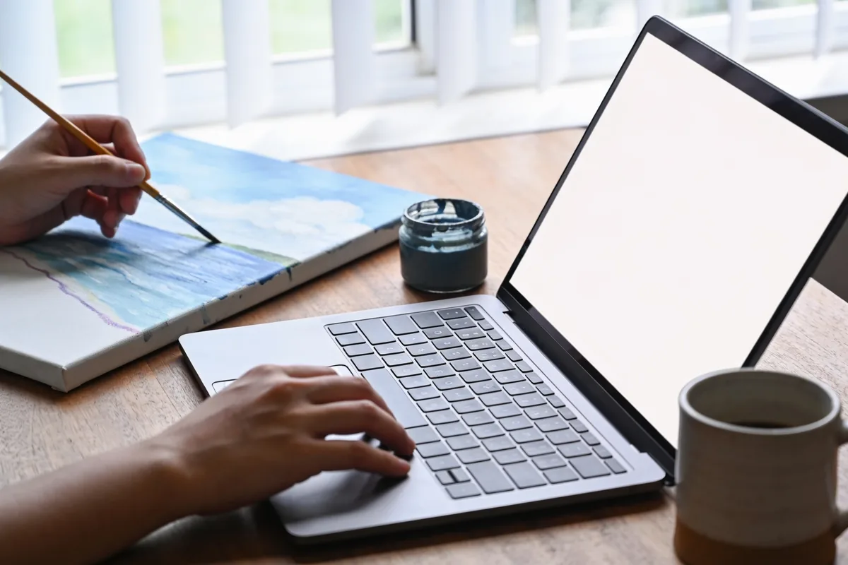 A person's hands holding a paintbrush over a canvas at a desk in a bright Australian home studio, with the other hand resting on a laptop keyboard, a pot of paint and a coffee mug nearby.