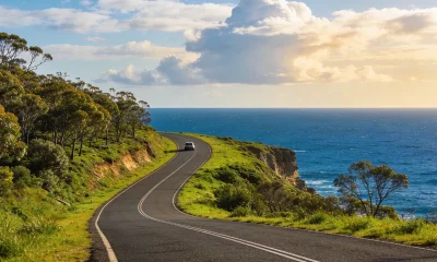 A sealed coastal road curving along a green clifftop high above the deep blue Southern Ocean on the Great Ocean Road in Victoria, with eucalypt bushland on the inland side and a single car in the middle distance under a big sky.
