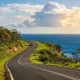 A sealed coastal road curving along a green clifftop high above the deep blue Southern Ocean on the Great Ocean Road in Victoria, with eucalypt bushland on the inland side and a single car in the middle distance under a big sky.