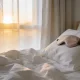 An unmade bed with rumpled white linen and a sleep mask resting on the pillow in a calm Australian bedroom, lit by soft morning sun glowing through sheer curtains.