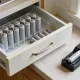 An open kitchen drawer in an Australian home holding plain unmarked rechargeable batteries upright in a clear organiser box with loose single-use batteries beside them, and a simple battery charger on the bench next to the drawer.
