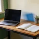 An open laptop and a notebook on a wooden home office desk by a window, with a mobile phone, a cup of tea and a small potted plant in soft morning light.