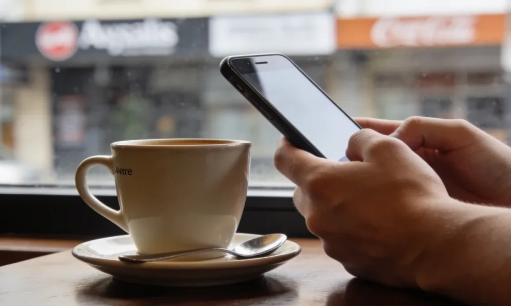 A person's hands holding a smartphone at a cafe table with a plain coffee cup beside it, the phone screen angled away from the camera in soft daylight.