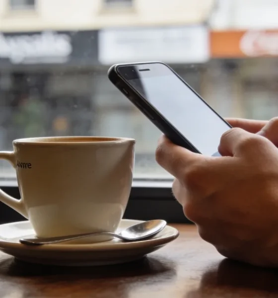 A person's hands holding a smartphone at a cafe table with a plain coffee cup beside it, the phone screen angled away from the camera in soft daylight.