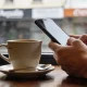 A person's hands holding a smartphone at a cafe table with a plain coffee cup beside it, the phone screen angled away from the camera in soft daylight.