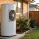 A plain white heat pump hot water unit standing on a concrete pad against the external wall of an Australian suburban house in soft morning light.