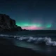 A faint green and pink aurora australis glow low over the southern ocean horizon from a dark Tasmanian beach at night, with a rocky headland silhouetted against the sky.