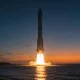 A large plain rocket lifting off from a coastal launch pad at dusk, its engine plume reflecting on calm water under a deep blue and orange twilight sky.