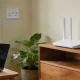 A plain white wi-fi router standing on a wooden shelf near a window in an Australian living room, with a laptop on a nearby table and a potted plant in natural daylight.