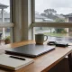 Timber home-office desk with a closed laptop, an open notebook and pen, a mug and a small external hard drive beside a rain-streaked window looking onto a wet suburban Australian street