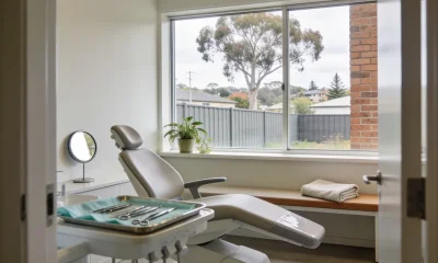 Calm dental surgery viewed from the doorway, with the dental chair angled towards a large window, a houseplant on the sill, and a gum tree, fence and brick wall beyond the glass