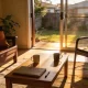 A bright, lived-in Australian living room in the late afternoon with two mugs, a closed folder and a pen on a low table, a chair pulled up to it and a sliding door open onto a small sunlit garden.