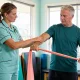 An allied health assistant guiding a client through a resistance band exercise in a bright therapy room.