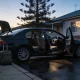 Dark sedan parked at the kerb on a quiet suburban Australian street at first light, its boot open and two soft-shell suitcases standing on the footpath beside it, both kerbside doors open