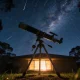 A backyard telescope on grass in an Australian suburb at night pointing at a starry sky with the Milky Way faintly visible, silhouetted gum trees and a warm-lit house window in the background.