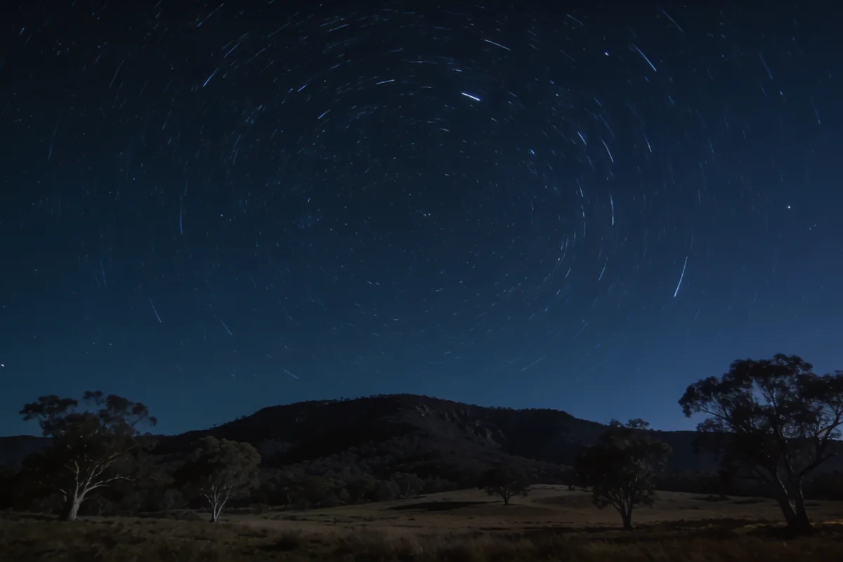 A long-exposure night sky over a quiet Australian landscape with faint star trails curving above dark hills and a few trees.