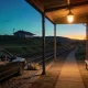 Dark sedan parked beside a country railway station platform at dusk, with a soft travel bag on the gravel behind it and a lit lamp over an empty timber bench, the station building glowing red brick