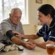 A community nurse in a navy uniform taking an older man's blood pressure at his kitchen table in a bright Australian family home.