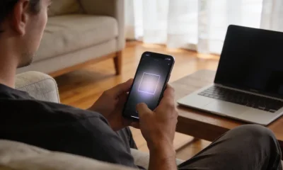 A person in a quiet living room viewed from behind over the shoulder, warily looking at a smartphone whose screen shows a softly blurred video frame, with a half-open laptop beside them.