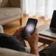 A person in a quiet living room viewed from behind over the shoulder, warily looking at a smartphone whose screen shows a softly blurred video frame, with a half-open laptop beside them.