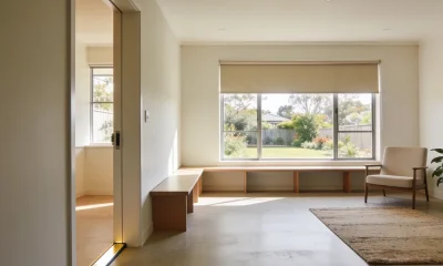 Spacious, ordinary Australian living area with a level doorway seen from the hallway, a long low bench under a large half-blinded window, an armchair and rug on a polished floor, and a garden beyond