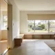 Spacious, ordinary Australian living area with a level doorway seen from the hallway, a long low bench under a large half-blinded window, an armchair and rug on a polished floor, and a garden beyond