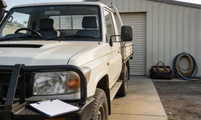 Right-hand-drive white work ute with a bull bar parked on the concrete apron of a small Australian business yard in clear morning light, a clipboard on the bull bar and a tool bag and coiled hose against a colourbond shed