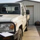 Right-hand-drive white work ute with a bull bar parked on the concrete apron of a small Australian business yard in clear morning light, a clipboard on the bull bar and a tool bag and coiled hose against a colourbond shed