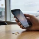A hand holding a smartphone above a light wooden cafe table with a small plastic SIM card lying beside it, an airport terminal and plane wing softly blurred beyond a window, the phone screen glowing with no readable content.