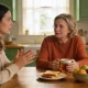 A mother and her adult daughter having a calm conversation at the kitchen table at home.