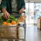 Shopper from behind filling a supermarket trolley with fresh fruit, vegetables, bread and milk in the produce aisle of a bright Australian grocery store