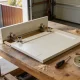 Cabinet door laid face down on a garage workbench with its two concealed cup hinges exposed, beside a screwdriver, tape measure, a saucer of screws, a mug and sawdust