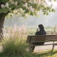 A person sitting on a park bench in an Australian spring park, seen from behind holding a tissue to their nose, a flowering tree and tall grass in bloom nearby in soft morning light.