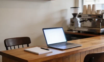 A tidy wooden counter in a small Australian shop or cafe with a blank notepad and pen, an open laptop on a blank glowing screen, and a second chair pulled up ready for a new team member, with bare walls behind.