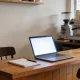 A tidy wooden counter in a small Australian shop or cafe with a blank notepad and pen, an open laptop on a blank glowing screen, and a second chair pulled up ready for a new team member, with bare walls behind.
