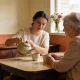 A home care worker pouring tea from a ceramic teapot for an older woman at a kitchen table in a warm Australian home.
