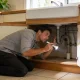 A homeowner kneeling to inspect the pipes under a kitchen sink with a flashlight in the morning light.