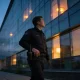 Security guard in a plain dark uniform on evening patrol outside a modern glass office building in Melbourne at dusk.