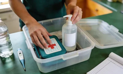 Hands packing a household emergency kit on an Australian kitchen bench with first-aid kit, hand sanitiser, water bottle and thermometer.