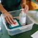 Hands packing a household emergency kit on an Australian kitchen bench with first-aid kit, hand sanitiser, water bottle and thermometer.