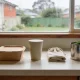 Brown paper takeaway container, a matte plant-fibre cup and a folded cloth produce bag in a row on a pale kitchen benchtop with a steel food-scrap caddy beside them, looking onto a rain-streaked window and a wet suburban garden