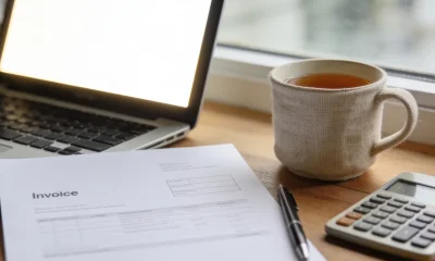 A small-business owner's desk by a window with an open laptop on a blank glowing screen, a plain white invoice page with no visible writing, a pen, a calculator and a cup of tea in soft natural light.
