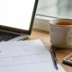 A small-business owner's desk by a window with an open laptop on a blank glowing screen, a plain white invoice page with no visible writing, a pen, a calculator and a cup of tea in soft natural light.