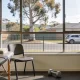Bright rehabilitation gym room with a treatment bed and folded towel beside a chair, a dumbbell and exercise step, and a large window onto a suburban Australian street with a gum tree